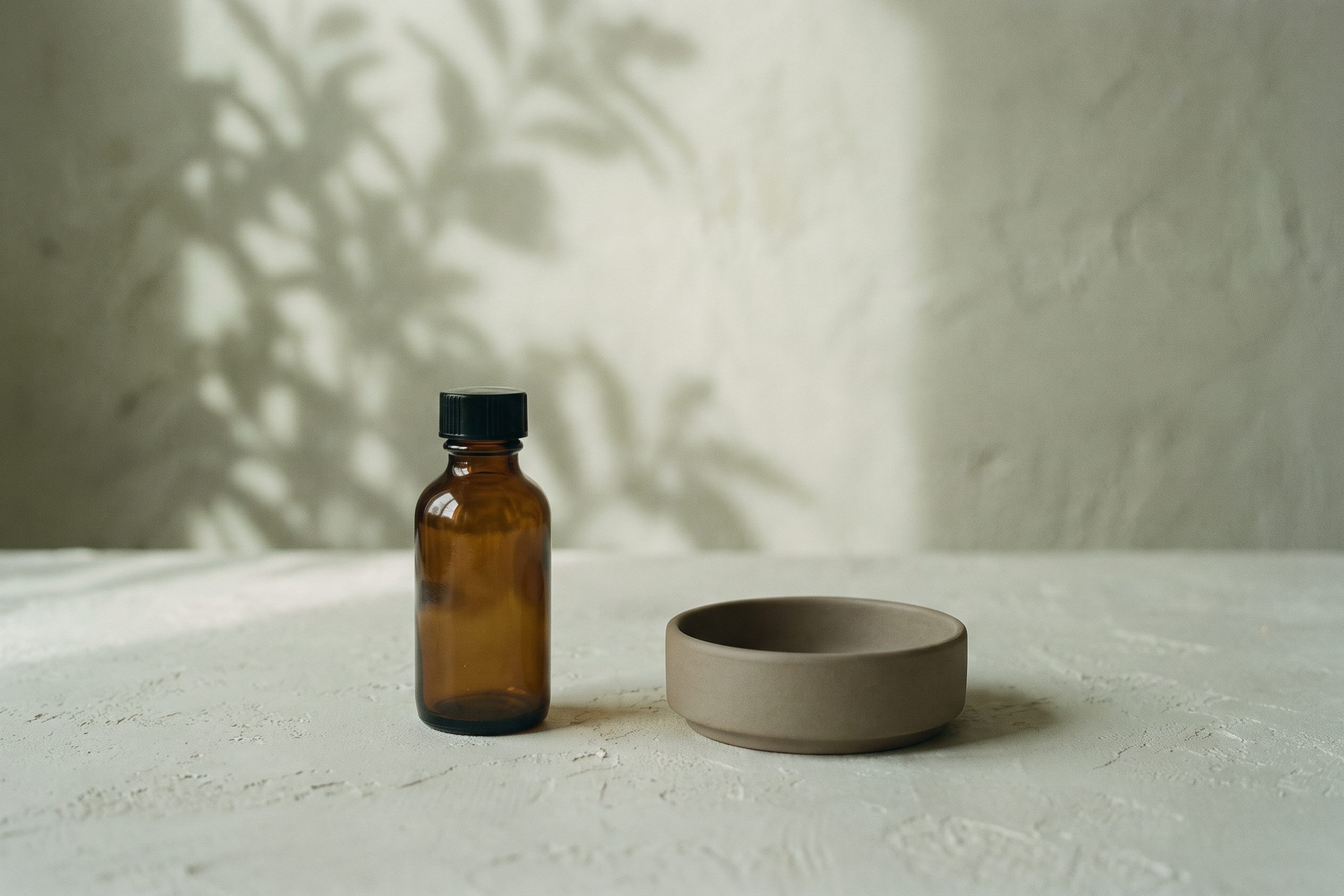 A small amber bottle and ceramic dish on plaster with a leaf shadow behind